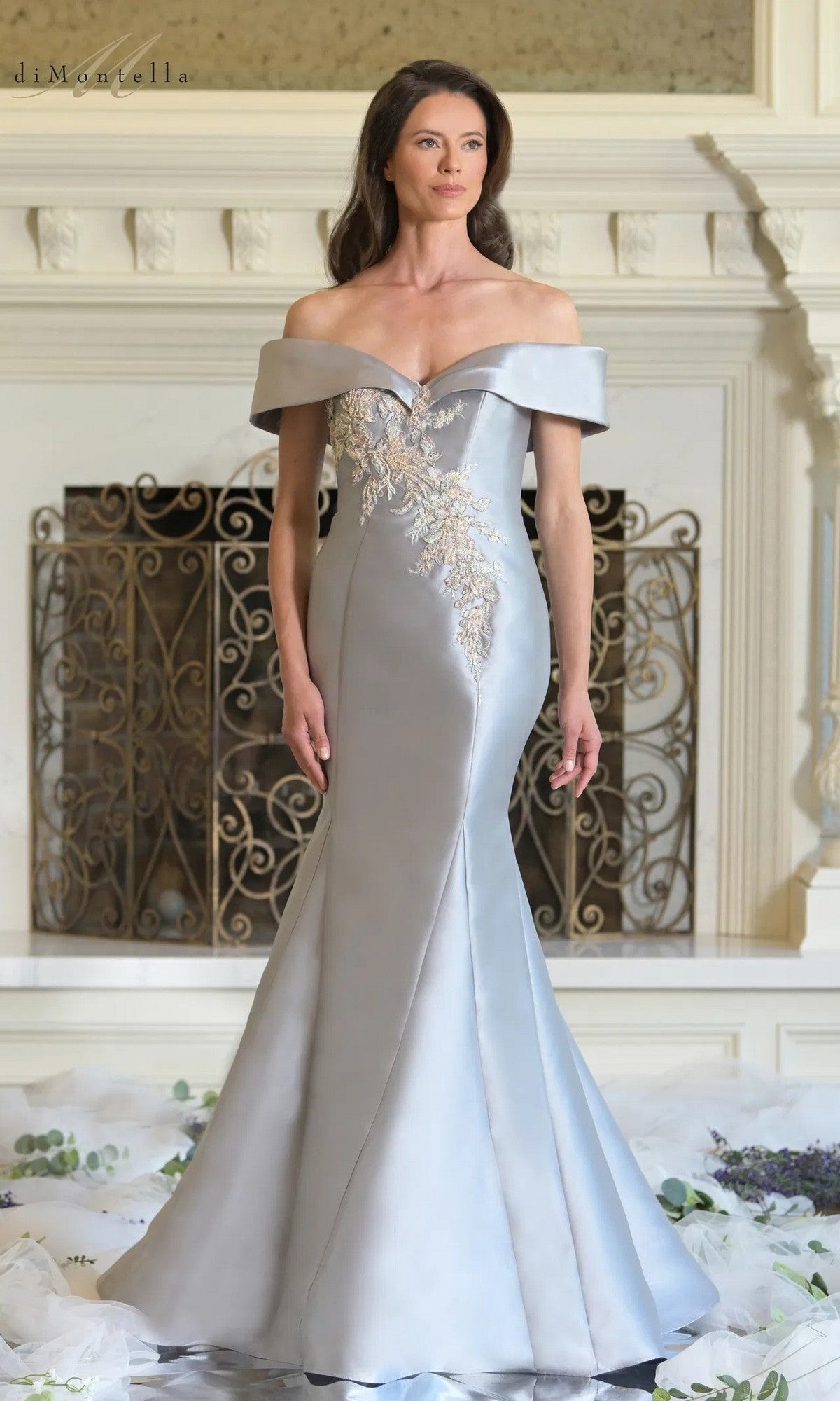 A woman stands indoors before an ornate fireplace, wearing the Long Formal Dress: Rina Di Montella RD3257 in platinum, featuring an off-the-shoulder design with beaded floral embroidery on the bodice.